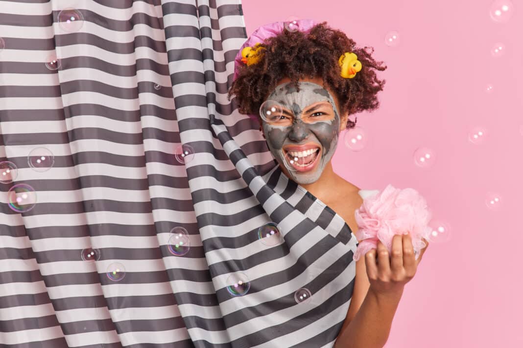 Annoyed curly haired woman frowns face exclaims loudly applies clay mask for skin treatment takes sh