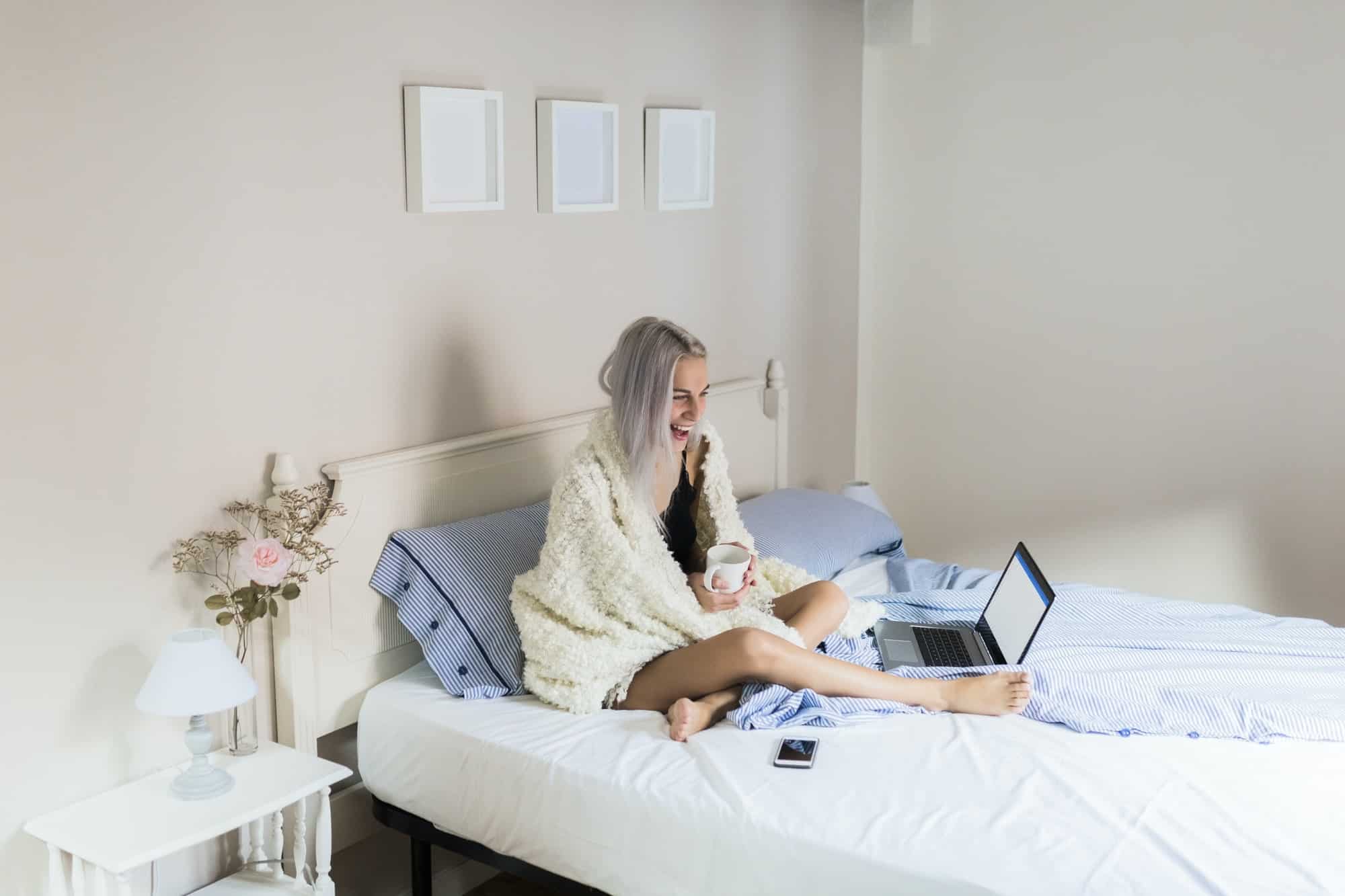 Happy young woman in bed with cup of coffee and laptop