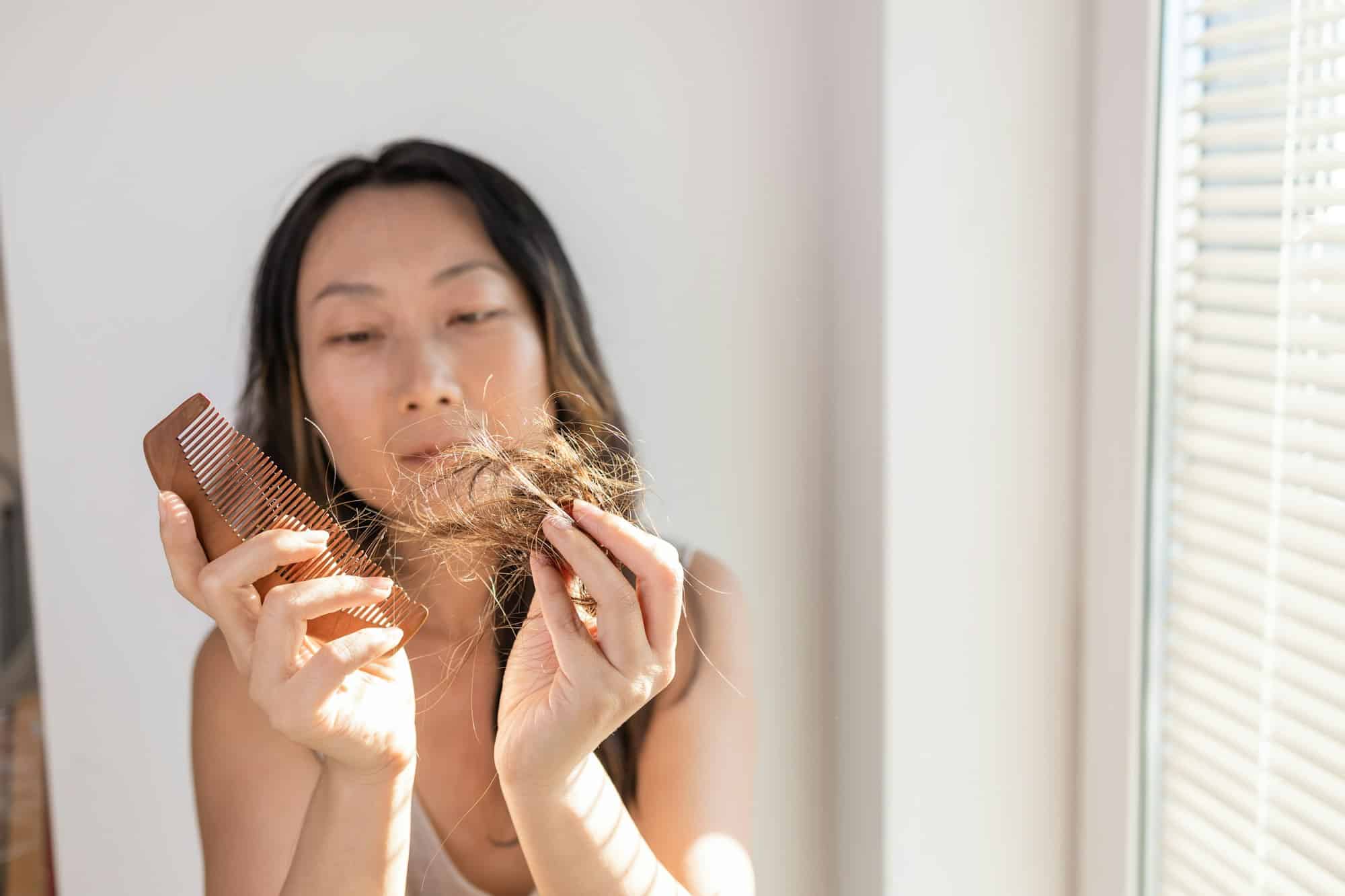 Young brunette girl holds a comb with hair falling out in her hand, looks worried and frustrated.